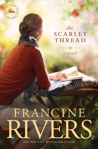A woman in a red shirt sits at a table outdoors reading a book.