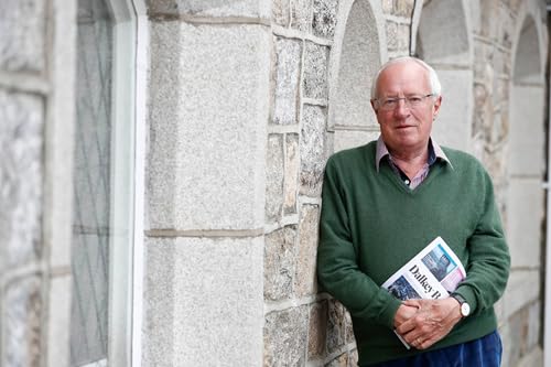 Older man in glasses holding a book against a stone wall.