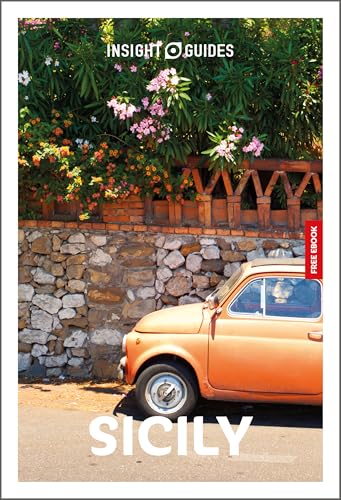 An orange vintage car parked in front of a stone wall with flowering bushes.