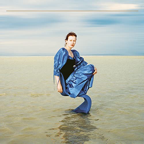 Woman in blue dress standing in shallow water, with cloudy sky in the background.