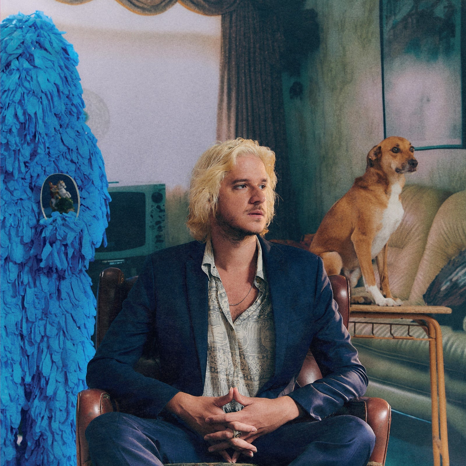 Blond man in suit sits with dog and blue sculpture.