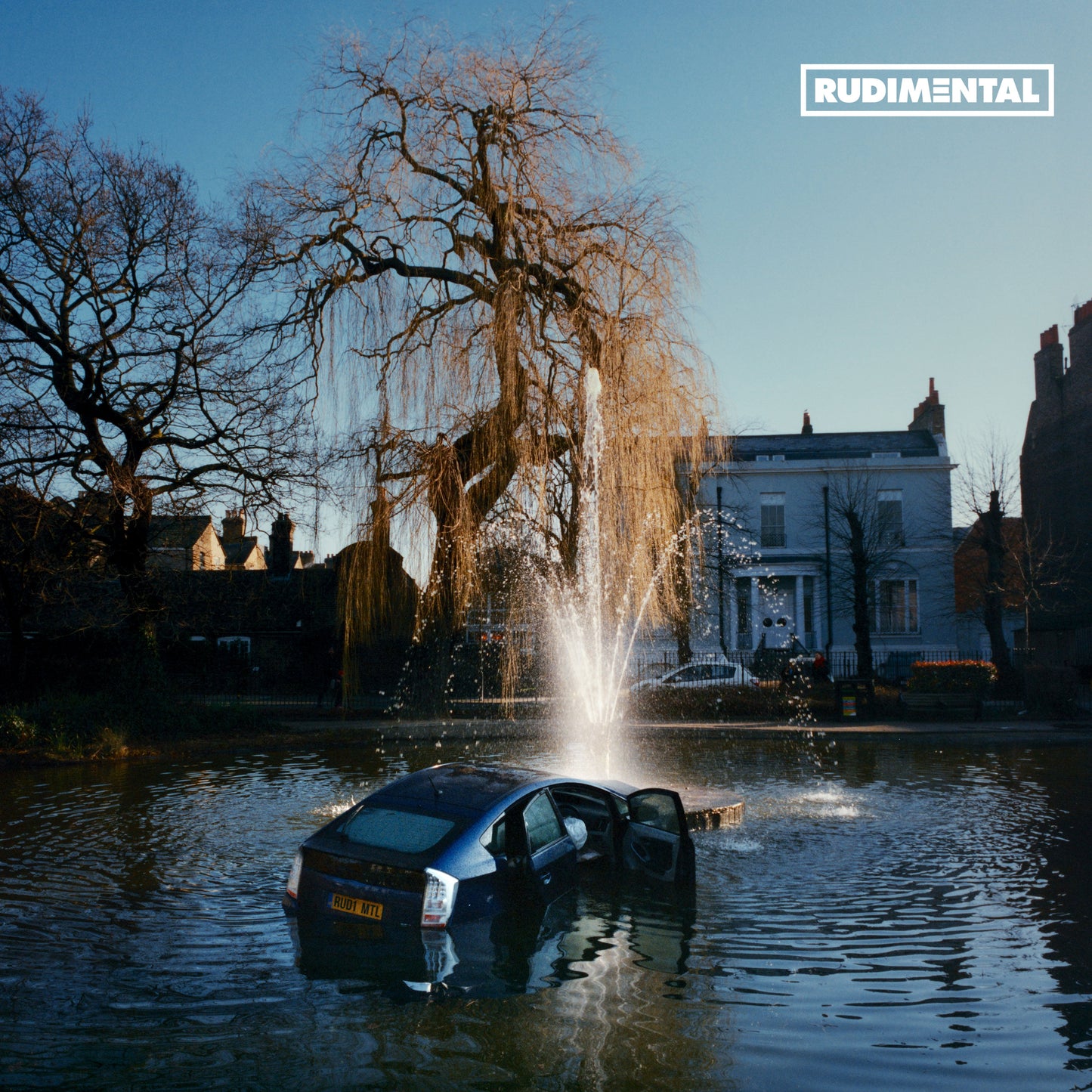 A blue car partially submerged in a park pond with a fountain spraying water.