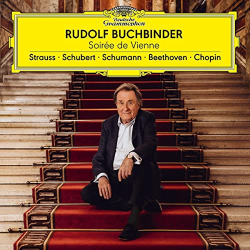 Rudolf Buchbinder sits on red stairs in a grand, ornate hall.