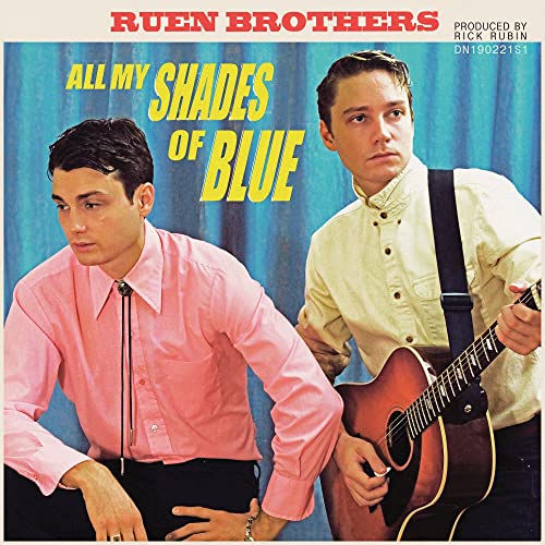 Two men posing in front of a blue curtain, one playing an acoustic guitar.