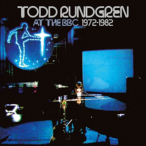 Todd Rundgren playing piano on stage with a blue light-up logo in the background.