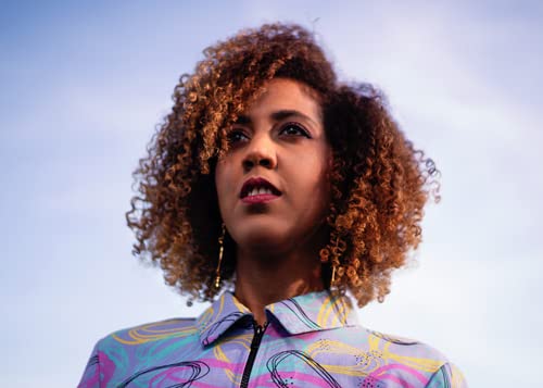 A woman with curly brown hair wearing a patterned shirt against a blue sky.