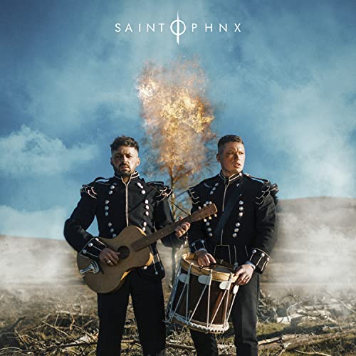 Two musicians in military-style jackets stand in a field with a burning tree.