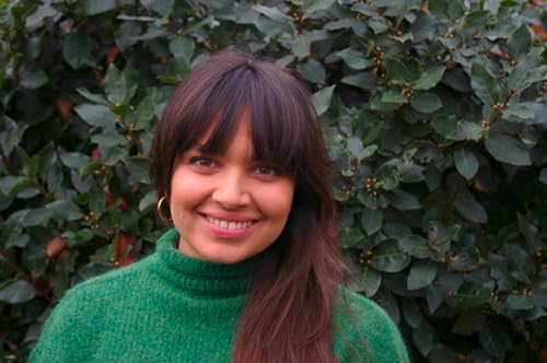 Woman with brown hair smiling, standing in front of green foliage.