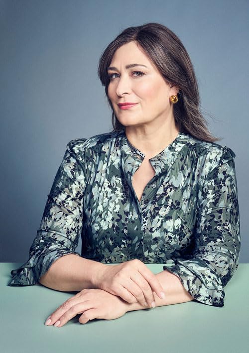 A middle-aged woman with brown hair wearing a green patterned blouse sits at table.
