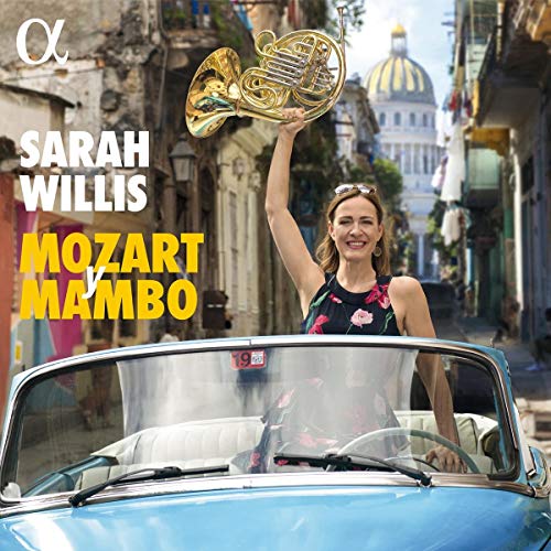 A woman stands in a blue convertible, holding a French horn aloft.