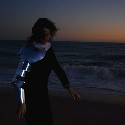 Woman in dark dress and partial armor stands on a beach.