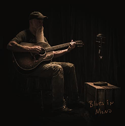 Seasick Steve sits playing an acoustic guitar with a vintage microphone.
