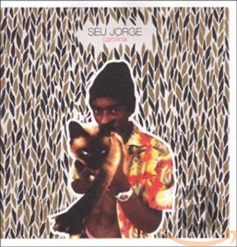 Seu Jorge wearing a beanie and floral shirt, holding a cat against leaf pattern.