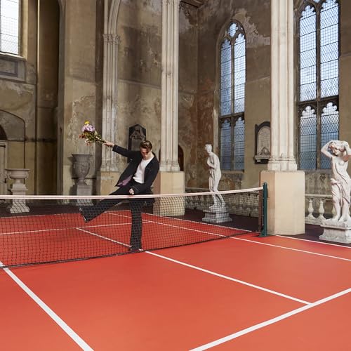 Man in suit poses on a red tennis court in a stone hall.