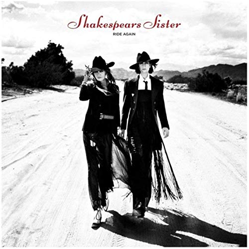 Two women in cowboy hats and dark suits walk down a dusty desert road.