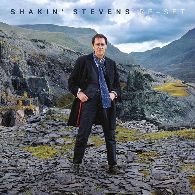 Shakin' Stevens stands in a rocky landscape wearing a dark coat and blue scarf.