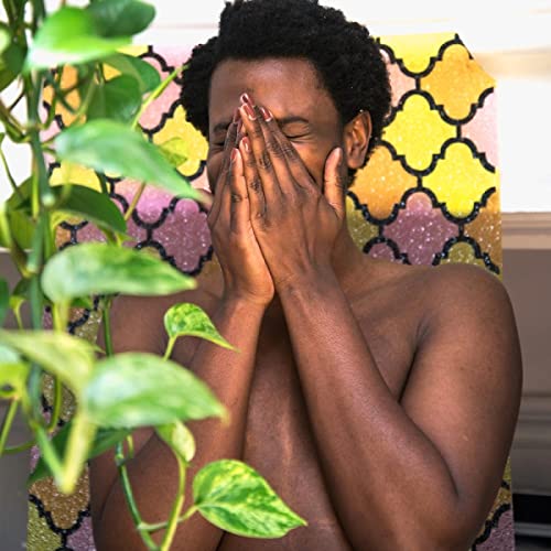Black man with eyes closed, covering face with hands, plants nearby.
