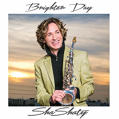A man with curly hair wearing a yellow patterned jacket holds a saxophone outdoors.