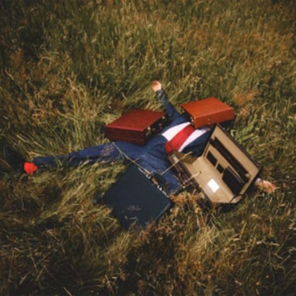 Person lying in grass surrounded by vintage suitcases.