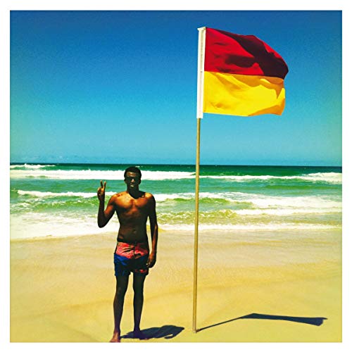 Shirtless man making a peace sign on a beach next to a red-yellow flag.
