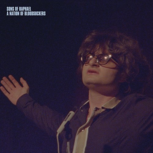 The image shows a man, possibly a musician, with glasses and outstretched hand.