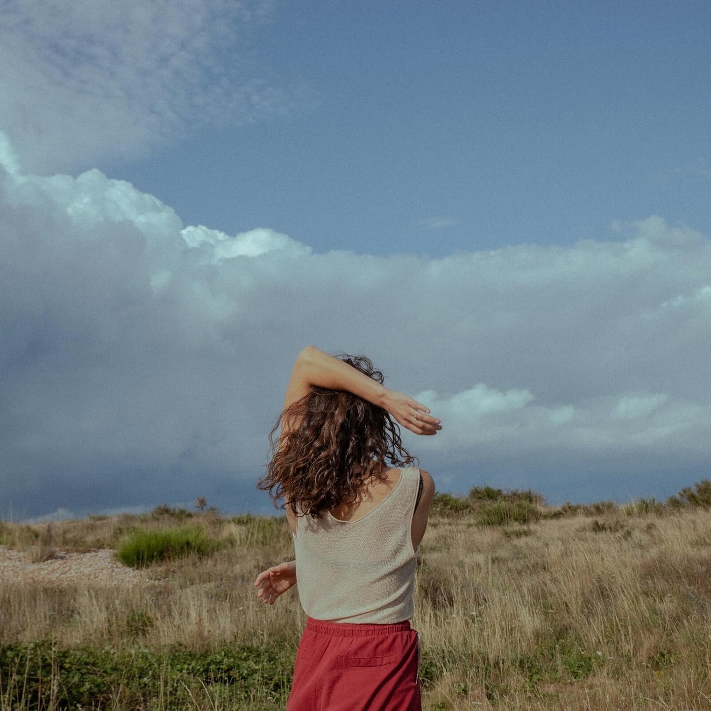 Woman with curly hair facing away, standing in a grassy field.
