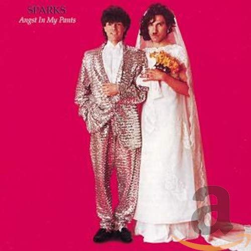 Two men stand against a pink background, one in a sequined suit, one in wedding dress.