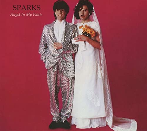 Two men stand against a red background, one in a sequined suit, one in bridal.
