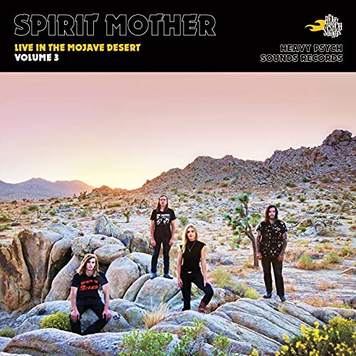 Four band members stand among large desert rocks at sunset under a black header.