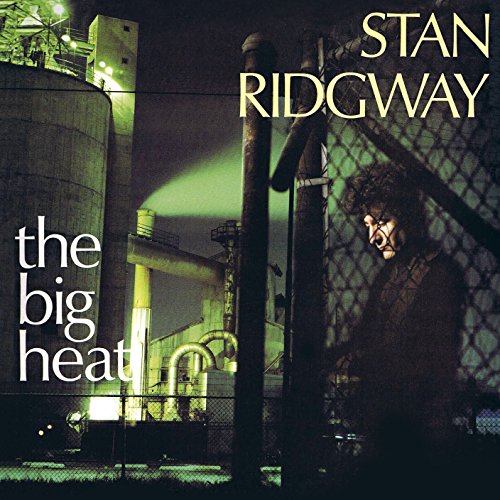 Stan Ridgway stands behind a chain-link fence at night near an industrial factory.