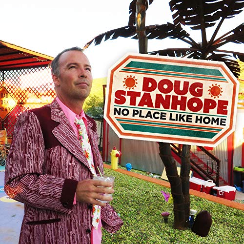 Doug Stanhope in a patterned suit holding a drink next to a sign.