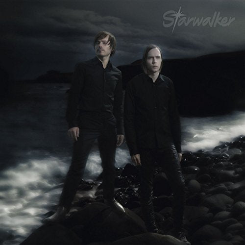 Two men in black stand on a rocky, misty shore.