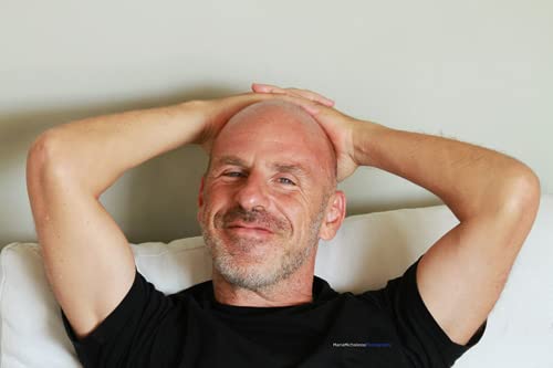 Bald man smiling with hands behind head, resting on a couch.