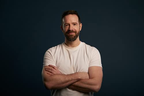 A man with a beard wearing a white t-shirt stands against a dark background.
