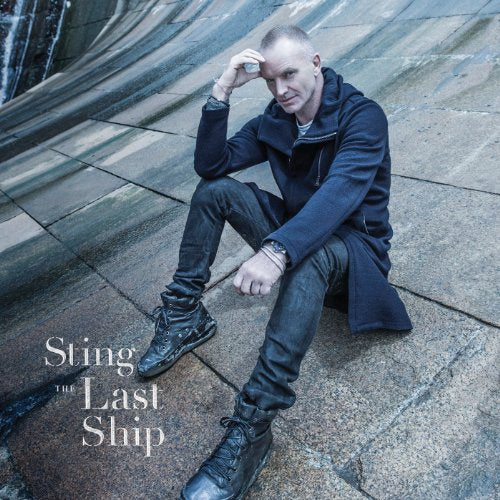 Sting sitting on a stone surface, wearing a dark jacket, jeans, and boots.