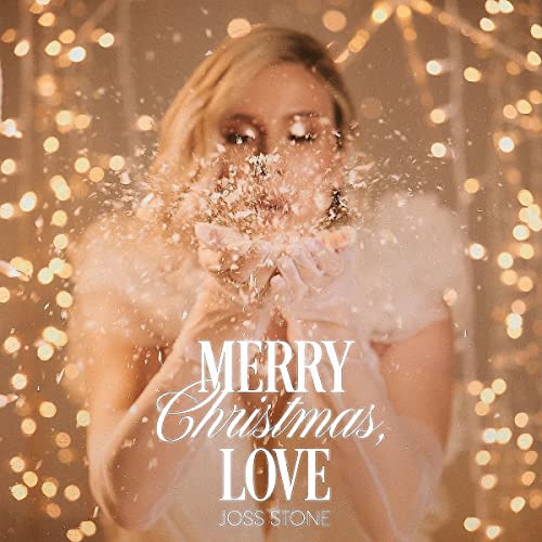 Joss Stone blows snow from her hands amidst a background of glowing golden fairy lights.
