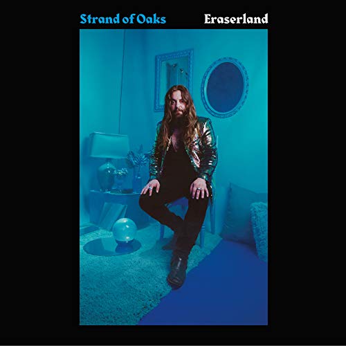 A long-haired man in a metallic jacket sits in a blue-tinted room.