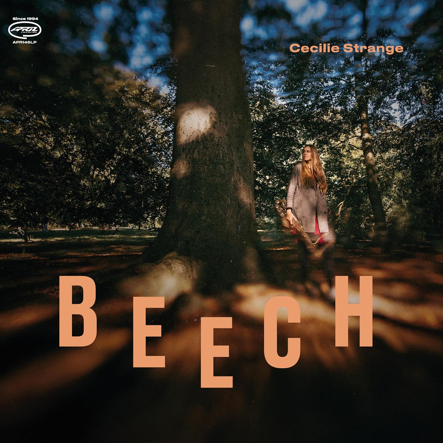 Cecilie Strange stands in a forest holding a saxophone next to a large tree.