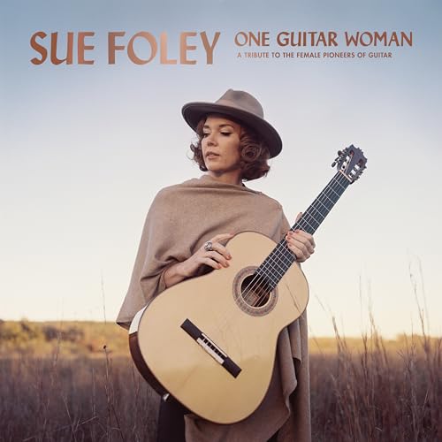 Sue Foley wearing a hat and poncho, holding an acoustic guitar in a field.