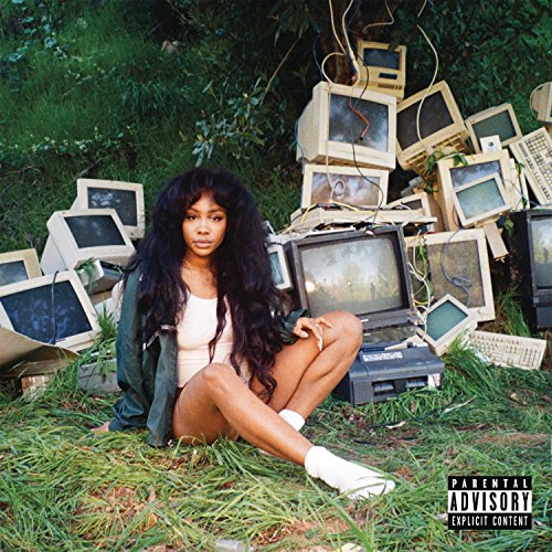 SZA sitting in grass surrounded by a pile of old computer monitors and keyboards.