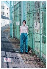 Young woman in casual clothes standing beside a green metal fence outdoors.