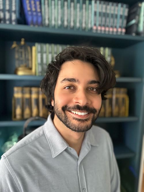 Man with dark hair and beard smiles, books in background.