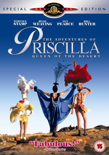 Three performers in elaborate drag costumes stand on a desert landscape against blue sky.