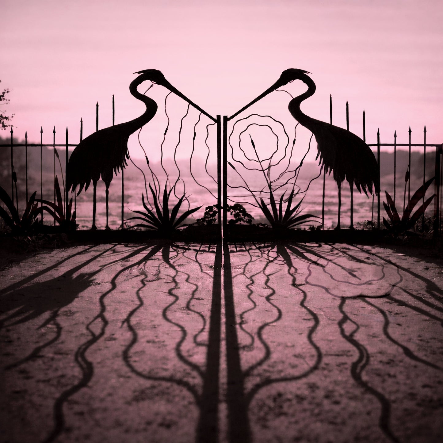 Silhouetted cranes flank a metal gate against a pink, hazy sky.