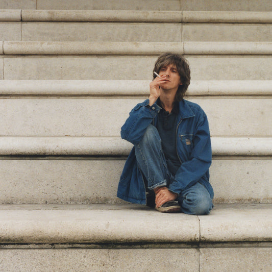 A man in a denim jacket sits on stone stairs smoking a cigarette.