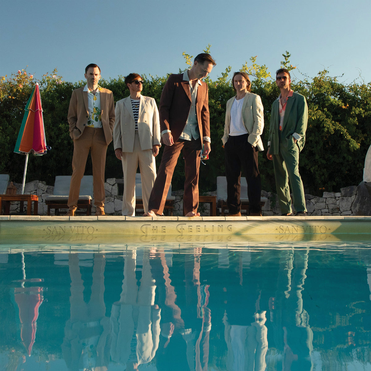 Five men in suits standing by a swimming pool, reflected in the water.