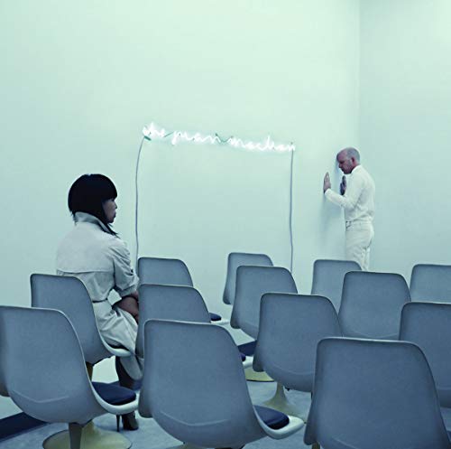 A woman sits in a room of chairs while a man faces the wall.