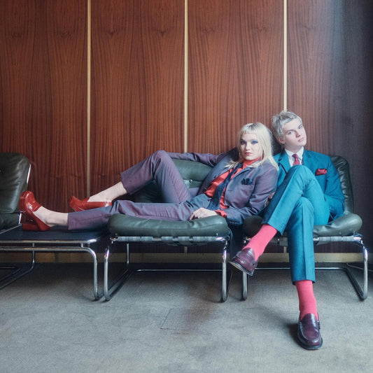 A man and woman in suits sit on chairs against a wood-paneled wall.
