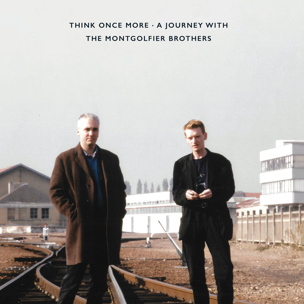 Two men stand on train tracks before a building, album cover.