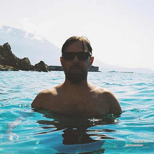 Man waist-deep in blue water, wearing sunglasses, mountains in background.